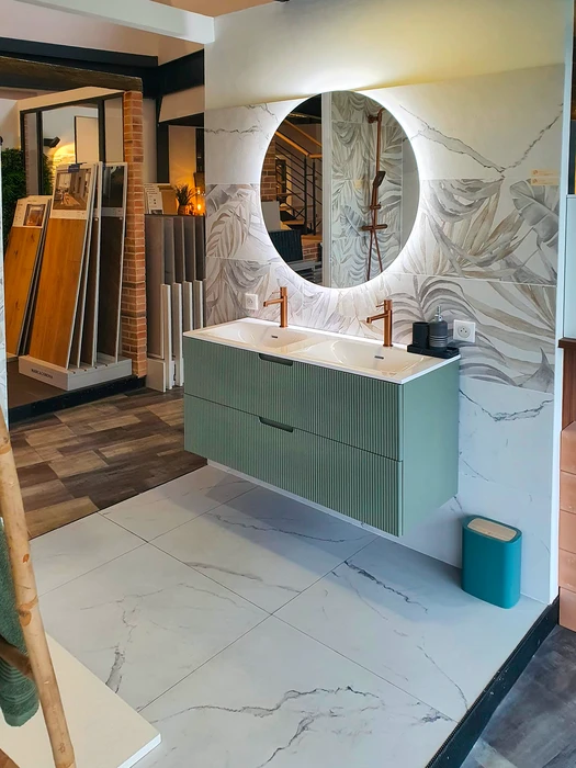 Showroom de salle de bain moderne avec meuble double vasque suspendu en vert sauge à façades striées, plan vasque blanc intégré, robinetterie cuivre brossé