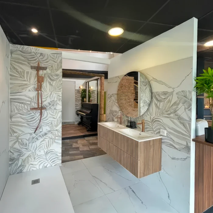 Showroom salle de bains moderne avec double vasque encastrée en marbre sur meuble bois cannelé suspendu, robinetterie cuivre brossé, miroir rond, carrelage grand format et revêtement mural décor tropical